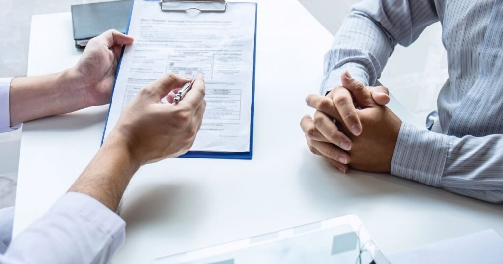 Person signs a contract on a clipboard while the other person sits with hands folded on a white table.