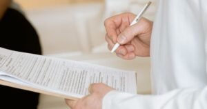 Person in a white coat holding a clipboard and writing on forms with a white pen
